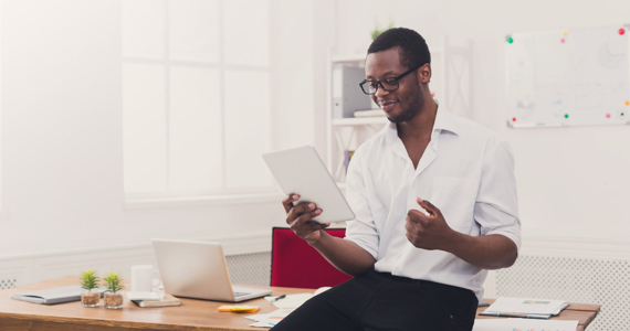 Jeune homme assis sur son bureau regarde une tablette en souriant