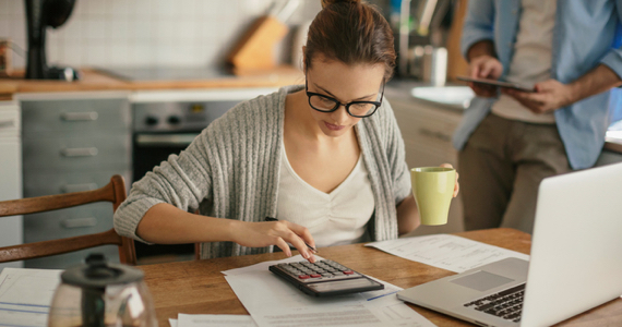 Jeune femme assise à sa table de cuisine fait ses comptes avec sa calculatrice