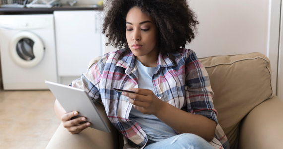 Jeune femme assise dans un fauteuil tient une tablette dans une main et une carte bancaire dans l’autre