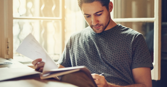 Jeune homme assis à un bureau devant une fenêtre lit un document en tenant un stylo