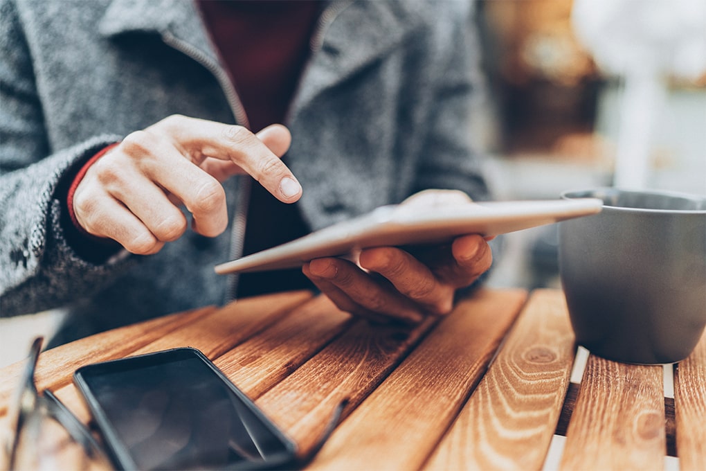 Image des mains d’un homme utilisant une tablette sur une table avec une tasse et un téléphone cellulaire.